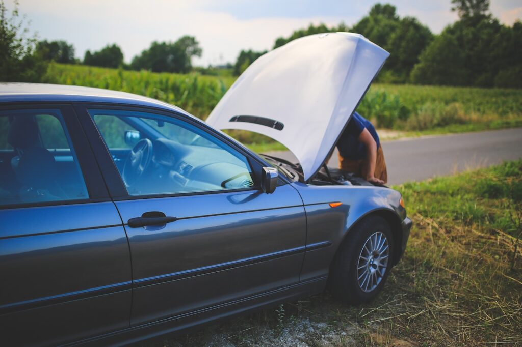 A man looking under the hood of his broken down car on the size of a country road.